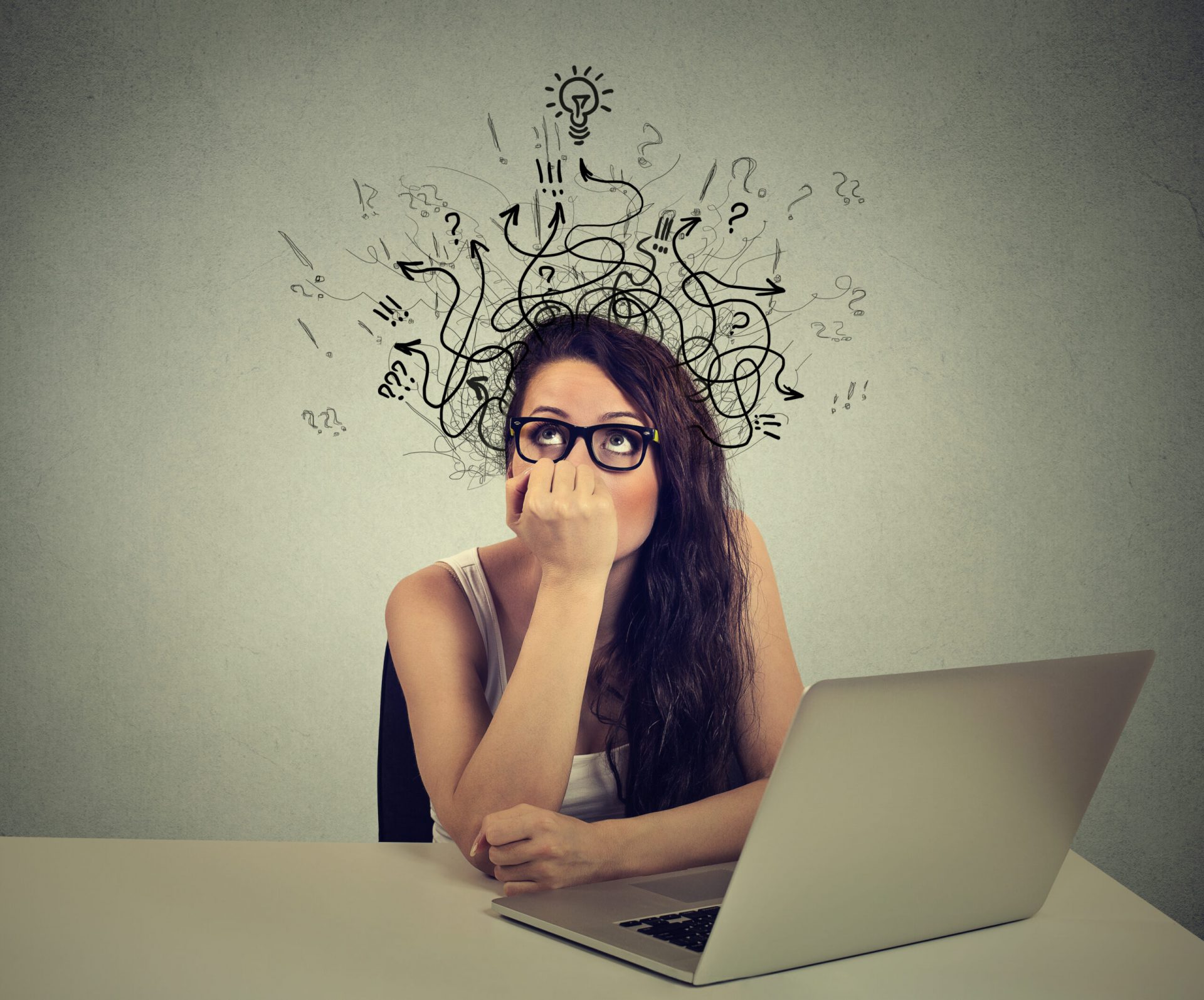 Young woman with thoughtful expression sitting at a desk with laptop with lines arrows and symbols coming out of her head